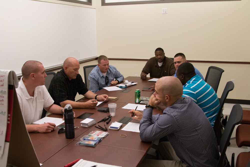 Seven men are seated around a large conference table in a meeting room, engaged in discussion. Papers, notebooks, and beverages are spread across the table, suggesting a structured work session or training meeting. A whiteboard or flip chart is partially visible on the left side of the frame.