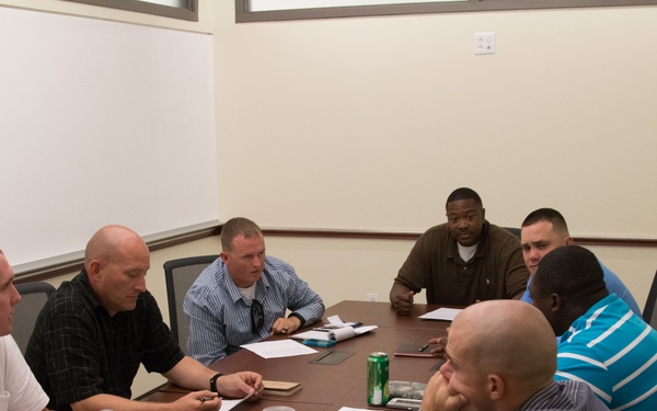 Seven men are seated around a large conference table in a meeting room, engaged in discussion. Papers, notebooks, and beverages are spread across the table, suggesting a structured work session or training meeting. A whiteboard or flip chart is partially visible on the left side of the frame.