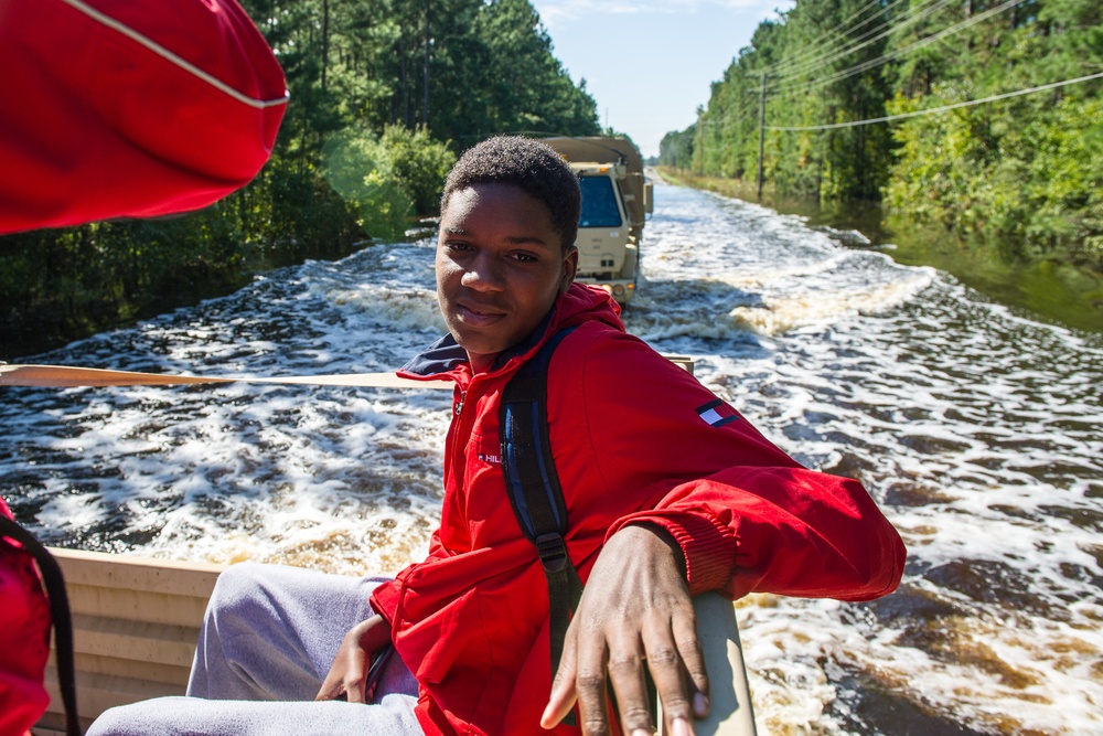 A young person wearing a red Tommy Hilfiger jacket sits in a flat-bottomed rescue boat traveling through a flooded road. A military-style truck is visible in the background also navigating the floodwaters. The scene appears to be an active flood rescue or evacuation operation, with trees lining both sides of the submerged roadway.