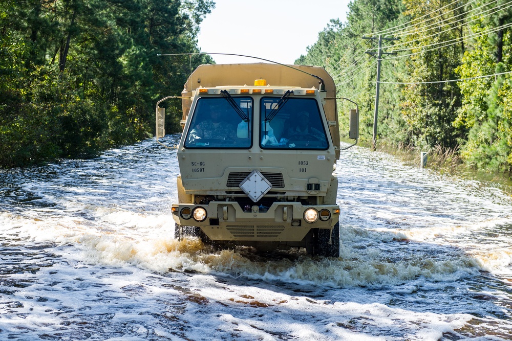 A tan/beige military tactical truck is driving through a flooded road surrounded by trees and utility lines. The vehicle appears to be a LMTV (Light Medium Tactical Vehicle) navigating floodwaters, likely during a disaster relief or emergency response operation. Water is visibly churning around the large tires as the truck pushes through the shallow but widespread flooding.