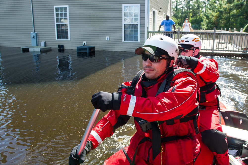 Flood rescue operation showing emergency responders in red dry suits and white helmets navigating a flooded residential area by boat. The floodwaters have submerged the ground level of nearby homes, with HVAC units and electrical panels partially underwater. Civilians are visible on an elevated deck in the background, suggesting they may be awaiting rescue or observing the operation.