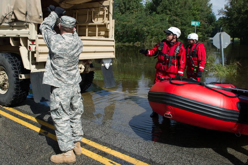 A flood rescue operation is underway on a partially submerged road. A U.S. Army soldier in ACU camouflage uniform interacts with two water rescue personnel in red drysuits and helmets standing beside a red inflatable rescue boat. The scene appears to be in a residential area with significant flood water covering the road and surrounding area.