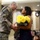 A U.S. Army soldier in ACU (Army Combat Uniform) is presenting a bouquet of yellow roses to a woman in a formal navy dress during what appears to be a military ceremony. The event is taking place in a military installation auditorium or meeting room decorated with unit insignia patches on the walls. An audience of soldiers and civilians, including children, is seated in the background observing the moment.