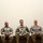 Three U.S. Army soldiers in Army Combat Uniform (ACU) are seated side by side in a formal or ceremonial setting, likely during an official military event or ceremony. All three are seated upright with hands folded in their laps, facing forward with composed expressions. The plain background and staged seating arrangement suggest a formal photo opportunity or panel setting.