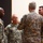 Four U.S. Army soldiers in Army Combat Uniform (ACU) digital camouflage pattern are participating in what appears to be a military decoration or promotion ceremony. A senior soldier facing away from the camera is pinning or presenting something gold-colored to a soldier identified by nametape as 'MORGAN'. Two other soldiers stand witness to the ceremony.