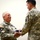 Two U.S. Army soldiers in ACU (Army Combat Uniform) digital camouflage are engaged in a formal military ceremony involving the transfer of a folded American flag. The soldier on the left, an older servicemember with gray hair and a mustache wearing a 1st Cavalry Division patch, is receiving or presenting the folded flag. A third soldier in U.S. Army uniform is visible in the background.