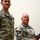 Two U.S. Army soldiers in ACU (Army Combat Uniform) digital camouflage are posed together indoors against a plain light-colored wall. The soldier on the right is holding what appears to be a rifle or long firearm. Both wear name tapes and unit patches, including U.S. flag patches on their left shoulders.