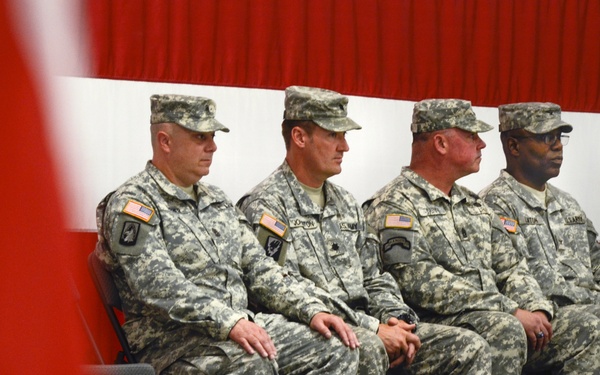 Four U.S. Army soldiers in Army Combat Uniform (ACU) are seated in a row on a raised stage or platform during what appears to be a formal military ceremony. All four wear patrol caps and desert tan combat boots, with American flag patches visible on their left shoulders. A red and white striped backdrop and a partially visible American flag in the foreground suggest a formal military event.