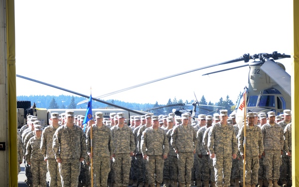 A large formation of U.S. Army soldiers in ACU (Army Combat Uniform) camouflage stands at attention outside a military hangar. The formation is viewed from inside the hangar, framed by the large hangar doors, with two soldiers positioned at each door panel. A CH-47 Chinook helicopter (tail number 102) is visible in the background among the assembled troops.