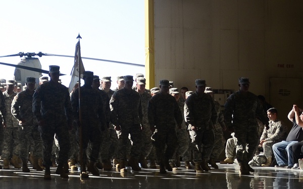 A large formation of military personnel in camouflage uniforms marches across a wet reflective tarmac or hangar floor, with a flag bearer visible among the ranks. Civilian spectators, including family members, are seated and standing along the right side observing the formation. The setting appears to be a military hangar or airfield, with at least one helicopter visible in the background.