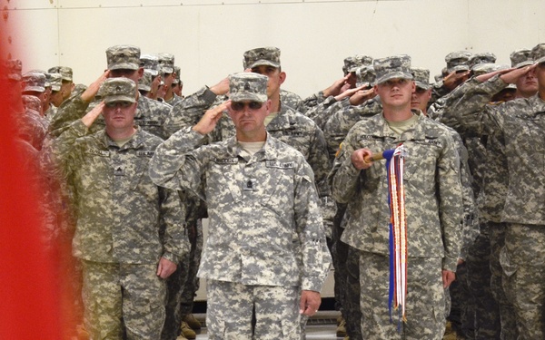 A large formation of U.S. Army soldiers in ACU (Army Combat Uniform) digital camouflage are standing at attention and saluting during a formal military ceremony held indoors, likely in a hangar or large facility. A red, white, and blue ribbon or streamer is visible being held by one soldier in the front row, suggesting a unit citation, activation, or deactivation ceremony. A U.S. flag is partially visible in the foreground left.