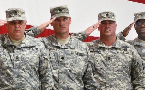 Four U.S. Army soldiers in ACU (Army Combat Uniform) digital camouflage are standing in a row and rendering a salute. They are positioned in front of a large American flag backdrop, suggesting a formal military ceremony or official event. Name tapes are partially visible on their uniforms, with readable names including 'Johnston' and 'Nunn'.