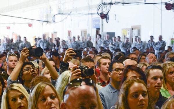 A large crowd of civilians and uniformed military personnel are gathered inside what appears to be a military hangar or large indoor facility. The civilians in the foreground are seated and attentively watching an event, while uniformed service members stand in formation in the background. Several attendees are holding cameras and photographing the proceedings, suggesting a formal military ceremony or event.