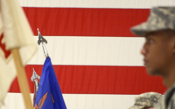 A U.S. Army military ceremony is taking place in front of a large American flag backdrop. Soldiers in Army Combat Uniforms (ACU) are participating in what appears to be a formal formation or change of command ceremony. Unit guidons, including a blue 'D' company guidon, are being carried by soldiers.
