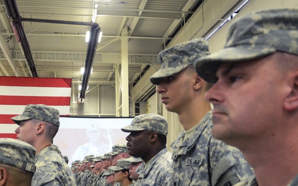 A formation of U.S. Army soldiers standing at attention inside a large indoor facility, likely a hangar or gymnasium. The soldiers are wearing Army Combat Uniforms (ACU) with digital camouflage pattern and patrol caps. A large American flag is displayed in the background alongside a projection screen, suggesting a formal military ceremony or briefing.