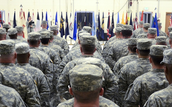 A large formation of U.S. Army soldiers in ACU (Army Combat Uniform) digital camouflage are assembled inside a military facility, facing toward a stage or podium area at the front. Multiple state and military flags are displayed along the back wall, suggesting a formal ceremony such as a deployment, reintegration, or unit activation event. Civilian attendees are visible on the left side, consistent with a family or community ceremony.