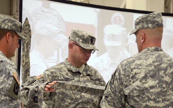 Three U.S. Army soldiers in ACU (Army Combat Uniform) digital camouflage are engaged in what appears to be a military ceremony, likely a casing or uncasing of colors. The central soldier is handling a folded guidon or flag while the others participate in the formal ritual. A projection screen displaying military imagery and a partial American flag are visible in the background.