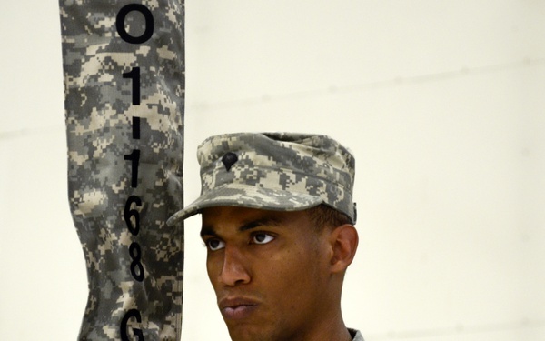 A U.S. Army soldier in ACU (Army Combat Uniform) digital camouflage stands at attention while holding a unit guidon or staff marked '0118 GSAB'. The soldier is the primary focus of the image, with at least two other uniformed soldiers visible in the background. The setting appears to be an indoor military formation or ceremony.