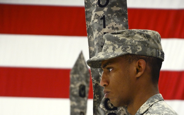 U.S. Army soldiers in Army Combat Uniform (ACU) digital camouflage are standing at attention during a formal military ceremony. A large American flag serves as the backdrop. The foreground soldier is holding a guidon or unit pennant with partial text visible reading 'CSAE' and the number '1'.