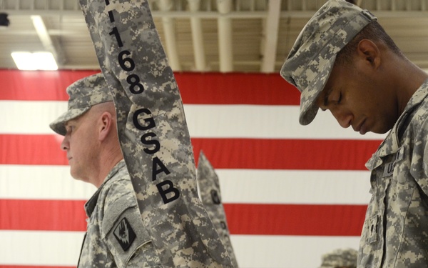 Two U.S. Army soldiers in ACU (Army Combat Uniform) digital camouflage are participating in a formal military ceremony, likely a change of command or unit activation/deactivation. They are handling a guidon (unit flag) bearing the designation '168 GSAB' (General Support Aviation Battalion) in front of a large American flag. The indoor ceremony is taking place in what appears to be a military hangar or gymnasium.