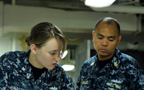 Two U.S. Navy personnel in Navy Working Uniform (NWU) are reviewing documents at a workstation aboard what appears to be a naval vessel. The female sailor on the left, identified by her name tape as 'DUNAGAN,' is examining a binder, while the male sailor on the right, whose name tape reads 'SHADD' and whose uniform displays 'U.S. NAVY,' points to a document. The setting appears to be an interior compartment of a ship with overhead piping and fluorescent lighting visible.