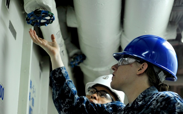 Two U.S. Navy personnel in Navy Working Uniform (NWU) camouflage are inspecting or operating a pipe valve in what appears to be a ship's interior compartment. The foreground sailor wears a blue hard hat and safety glasses, while the background sailor wears a white hard hat. The label 'FR100' is visible on the bulkhead, indicating a frame reference point aboard a naval vessel.