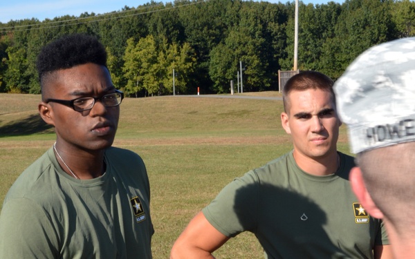 Three U.S. Army personnel are gathered outdoors on a grassy field, with two soldiers in olive green Army PT (Physical Training) shirts facing a third individual in ACU (Army Combat Uniform) whose back is to the camera. The two facing soldiers appear to be receiving instruction or feedback. The setting appears to be a military training area.