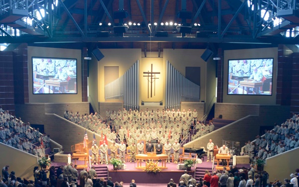 A large military ceremony or memorial service is taking place inside a spacious church or megachurch auditorium. The venue is filled to capacity with hundreds of attendees in civilian clothing in the main floor seating, while uniformed military personnel occupy the elevated choir loft and stage area at the front. Large projection screens on either side display the proceedings to the audience.