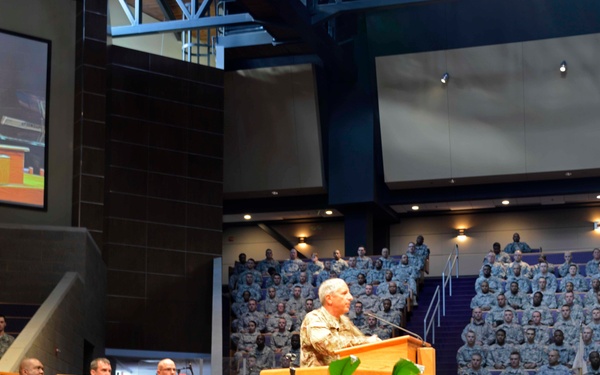 A military ceremony or formal address is taking place in a large auditorium or chapel, with a uniformed officer speaking at a wooden podium. The audience consists of hundreds of military personnel in camouflage uniforms filling tiered seating. A large projection screen on the left displays a live feed of the speaker at the podium.