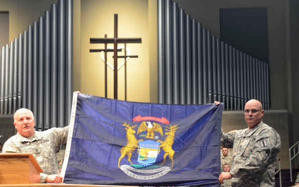 Two U.S. Army soldiers in ACU (Army Combat Uniform) camouflage are displaying the Michigan State flag during what appears to be a military ceremony held inside a chapel or church. The setting includes a pulpit, organ pipes, and a cross, suggesting this is a military chapel. An audience of uniformed personnel is seated in the background.