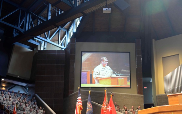 A military ceremony or formal address is taking place inside a large chapel or auditorium. A soldier in uniform is speaking at a podium on the right side of the stage, with his image projected on a large screen behind him. The audience consists of hundreds of uniformed military personnel seated in tiered seating.