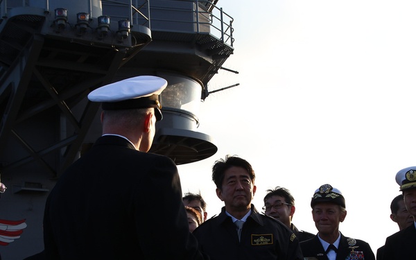 A formal military gathering on the deck of what appears to be a large naval vessel, likely an aircraft carrier, indicated by the superstructure and yellow 'E' marking visible in the background. A group of naval officers in dress uniforms, along with civilians, are assembled facing a senior officer whose back is to the camera. The setting suggests an official visit, briefing, or welcoming ceremony.