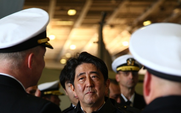 A civilian man in dark clothing is engaged in conversation with naval officers wearing white-topped dress uniform caps. The setting appears to be an indoor military facility, possibly the hangar deck of a naval vessel. Multiple uniformed personnel are visible in the background, suggesting an official visit or ceremony.