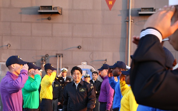 A dignitary in a brown jacket walks along a red carpet between two rows of personnel in colorful shirts who are rendering a salute. The setting appears to be the deck of a naval vessel, with wooden bollards with white rope lining the ceremonial path. Military officers in formal uniforms are visible in the background.