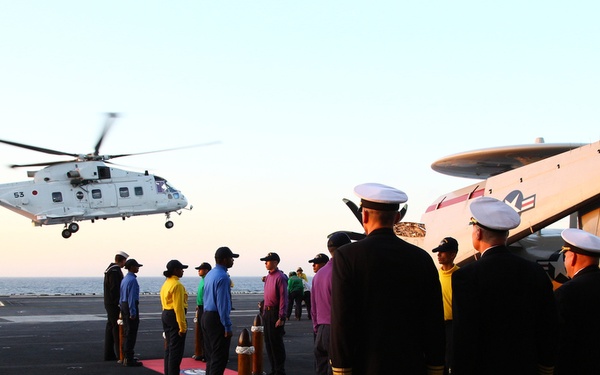 A military ceremony is taking place on the flight deck of an aircraft carrier, where naval officers in dress uniforms and deck crew in colored jerseys are lined up along a red carpet awaiting the arrival of a helicopter. The helicopter, marked with the number '53' and appearing to bear Japanese Maritime Self-Defense Force markings, is approaching to land. A fixed-wing aircraft, likely an E-2 Hawkeye (number 601), is visible parked on the deck in the background.