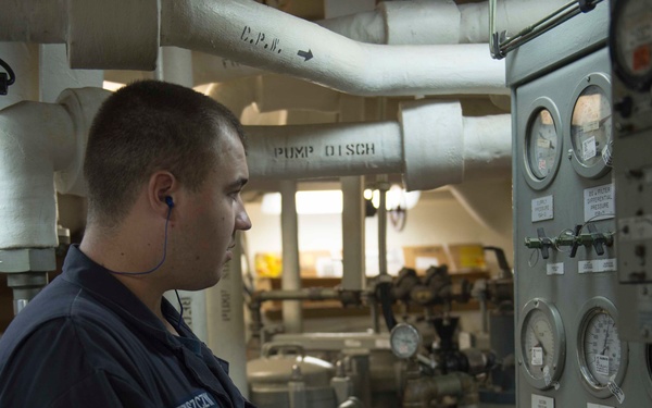 A uniformed personnel member, identifiable by a name tag reading 'STRYCZEK', is conducting an inspection or logging readings in an industrial machinery space, likely aboard a naval vessel. He is holding a clipboard with forms and wearing an earpiece while examining a panel of gauges and instruments. The environment features labeled pipes including 'PUMP DISCH' and a control panel with multiple pressure gauges.