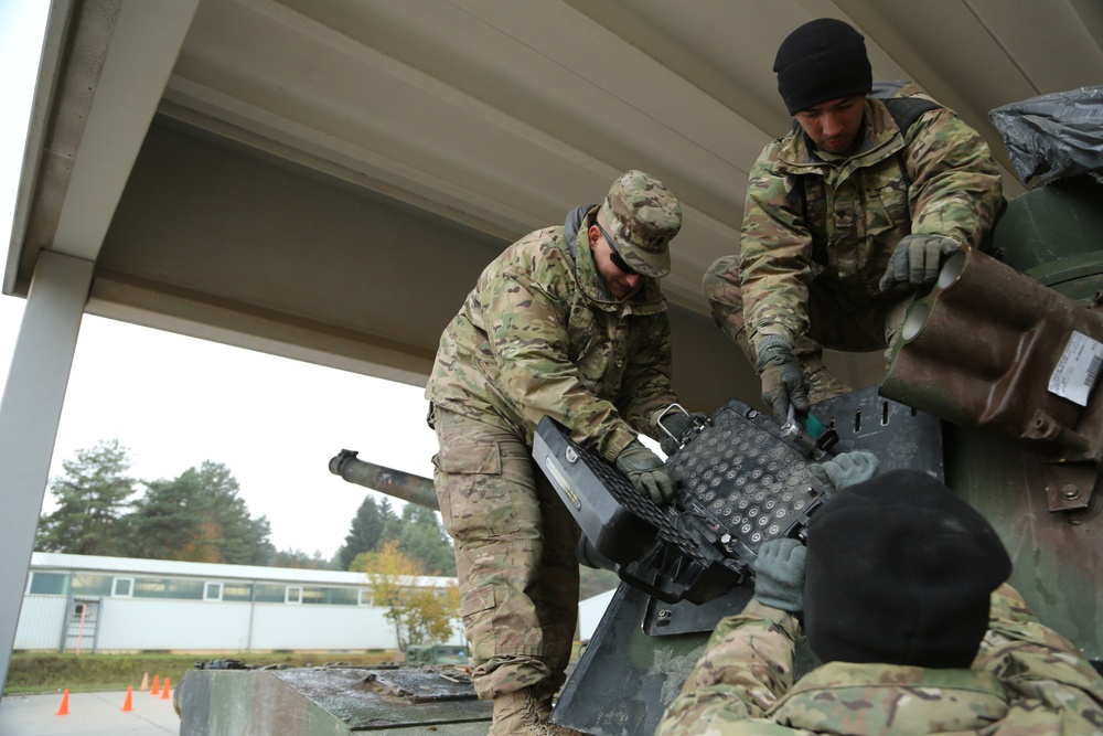 Three soldiers in MultiCam/OCP camouflage uniforms are working on top of a military armored vehicle, appearing to install or adjust equipment. The activity is taking place under a covered structure at what appears to be a military installation or base. The setting suggests a maintenance, equipment installation, or pre-deployment preparation task.
