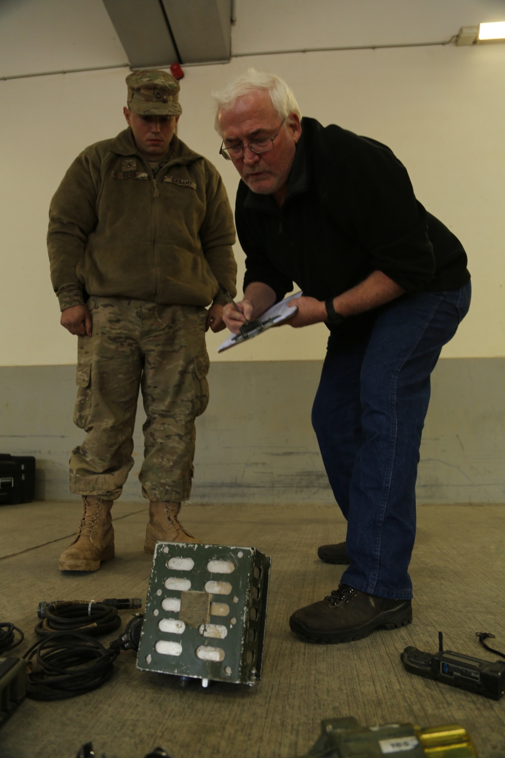 Two individuals are examining military equipment on a concrete floor in what appears to be an indoor facility or warehouse. A civilian in a black shirt and jeans is leaning over and writing on a clipboard, while a uniformed military personnel in MultiCam/OCP camouflage stands nearby observing. Several pieces of equipment, cables, and electronic devices are laid out on the floor for inspection or inventory.
