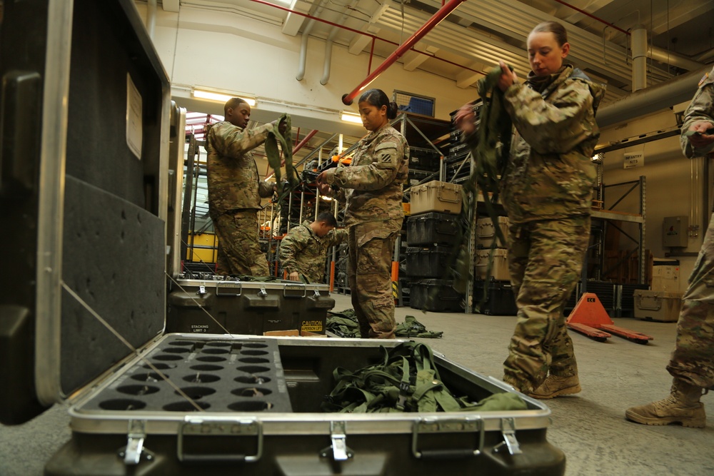 Military personnel in MultiCam/OCP camouflage uniforms are working inside a warehouse or supply depot, handling and organizing equipment. At least four soldiers are visible, engaged in what appears to be an inventory, packing, or equipment inspection operation. The setting contains military storage cases, shelving units, and various pieces of gear.