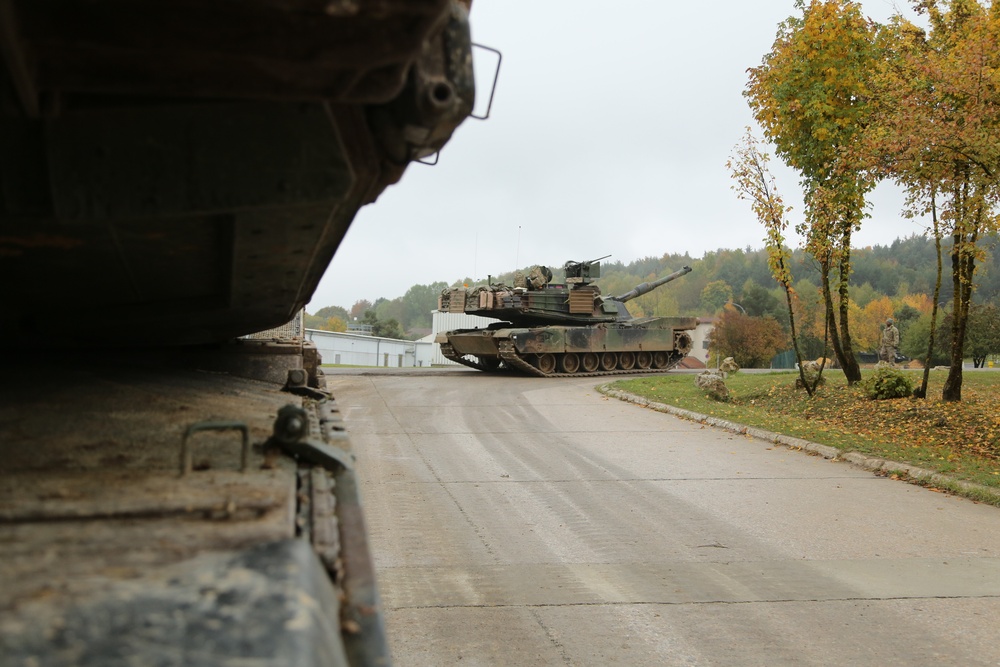 A military main battle tank, identifiable as an M1 Abrams, is maneuvering along a paved road within what appears to be a military installation or training base. The photo is taken from a low angle behind another armored vehicle in the foreground, with autumn trees and military personnel visible in the background. The setting suggests a training exercise or unit movement at a garrison facility.