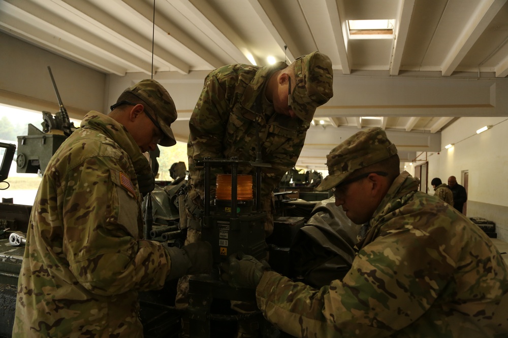 Three U.S. Army soldiers in MultiCam/OCP camouflage uniforms are working together on a piece of military equipment inside what appears to be a maintenance bay or motor pool facility. The soldiers are focused on inspecting or repairing a component that has an illuminated orange/amber light visible. Additional military vehicles and personnel are visible in the background.