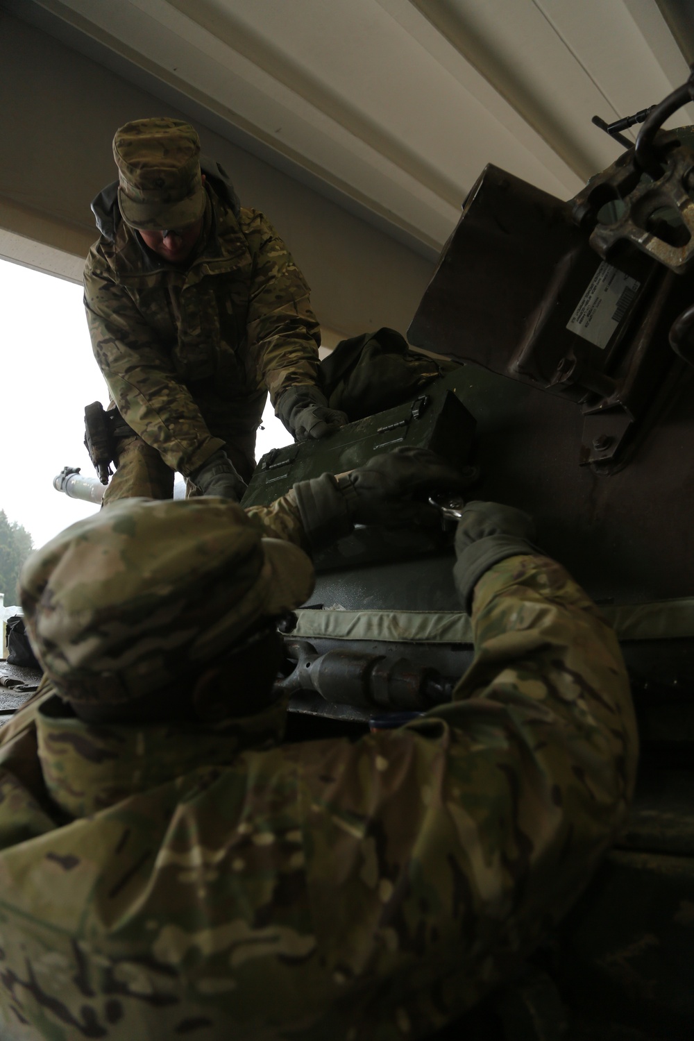Two military personnel in MultiCam/OCP camouflage uniforms are handling military equipment, appearing to load or transfer green metal ammunition cans or equipment cases. The image is taken from a low angle looking upward, with one soldier standing and leaning over while the other is positioned below. The setting appears to be under a covered structure or vehicle bay.