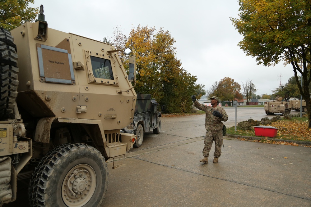 A soldier in camouflage uniform is directing or signaling near a column of military vehicles, including a tan MRAP (Mine-Resistant Ambush Protected vehicle) in the foreground. The scene appears to be at a military installation or staging area during autumn, with trees displaying fall foliage in the background. Additional military vehicles, including a Humvee and what appears to be a tracked vehicle with a red cross marking, are visible in the background.