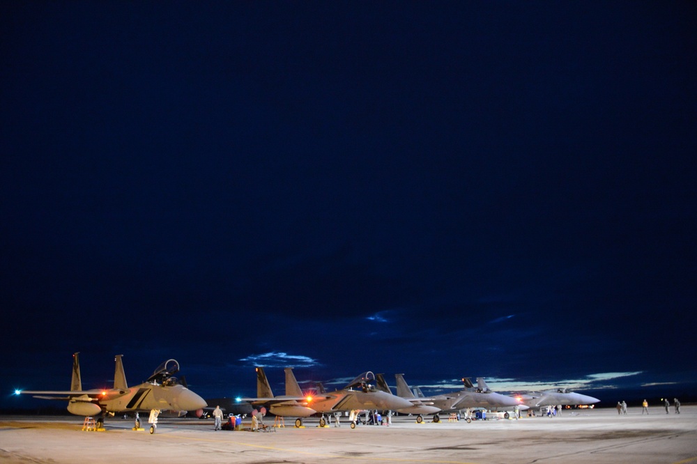 A nighttime flight line scene showing multiple military jet aircraft parked on a tarmac under a deep blue twilight sky. Ground crew personnel are visible working around the aircraft, illuminated by orange and white ground lighting. The scene suggests pre-flight or post-flight maintenance operations at a military air base.