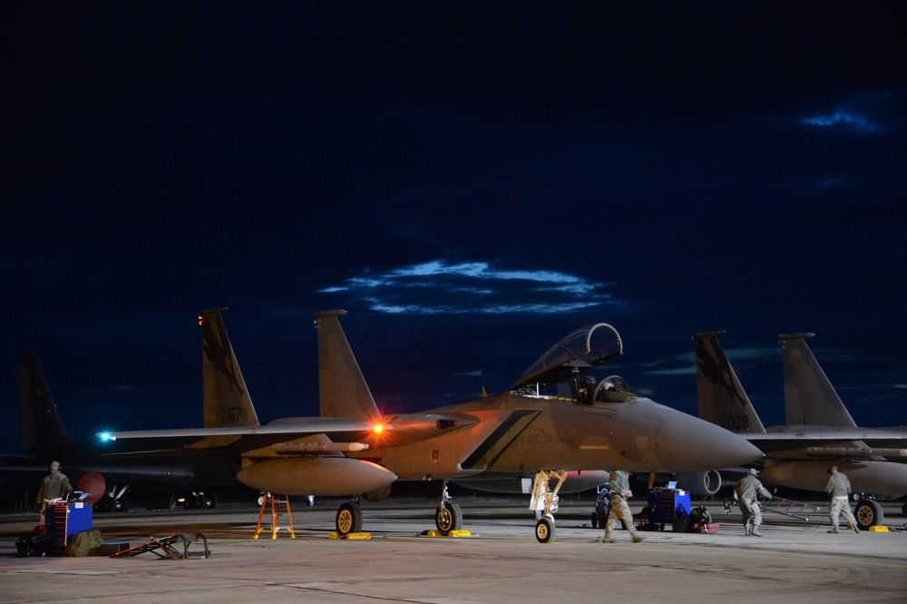 A nighttime flight line scene showing multiple F-15 Eagle fighter jets parked on an airfield tarmac. Ground crew personnel are conducting pre-flight or post-flight maintenance operations under artificial lighting. The dark sky with faint cloud formations provides a dramatic backdrop to the military aviation activity.