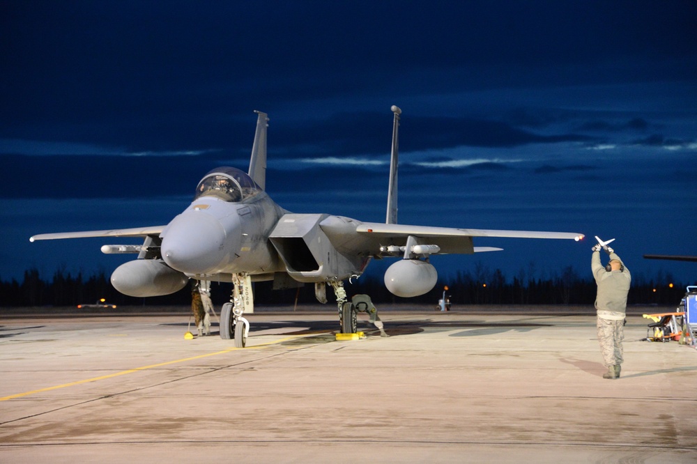 A U.S. Air Force F-15 Eagle fighter jet is parked on a flight line apron during twilight or early morning hours. Ground crew personnel are conducting pre-flight or post-flight maintenance checks around the aircraft. The scene is illuminated by artificial lighting against a deep blue sky.