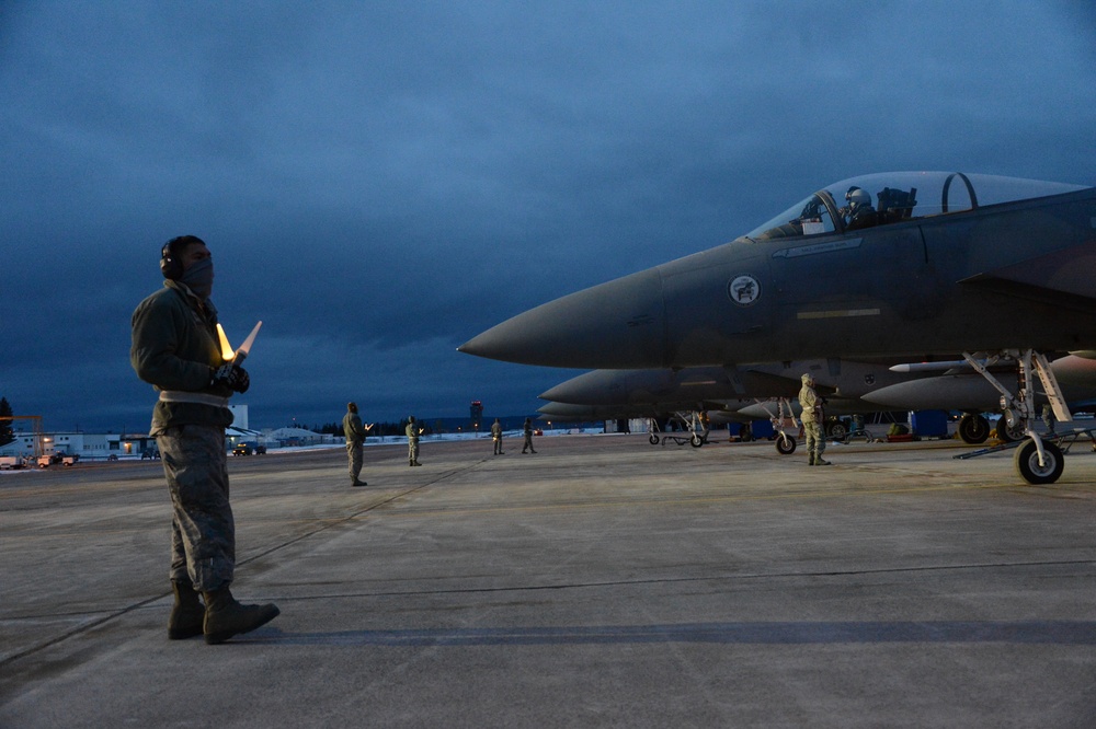 A military flight line scene captured at dusk or dawn showing ground crew personnel conducting pre-flight or post-flight operations around what appears to be an F-15 Eagle fighter jet. A crew chief in the foreground holds illuminated marshalling wands while a pilot is visible in the cockpit. Multiple ground personnel are positioned around the aircraft on the tarmac under an overcast sky.