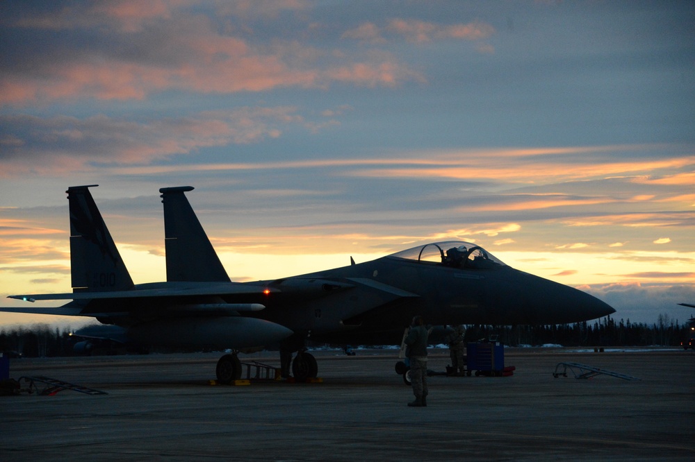 A military fighter jet, identifiable as an F-15 Eagle, is parked on a flight line apron during twilight or dawn conditions. At least one ground crew member is visible near the aircraft, likely performing pre-flight or post-flight maintenance checks. The dramatic sky with orange and pink cloud streaks provides a striking silhouetted backdrop.