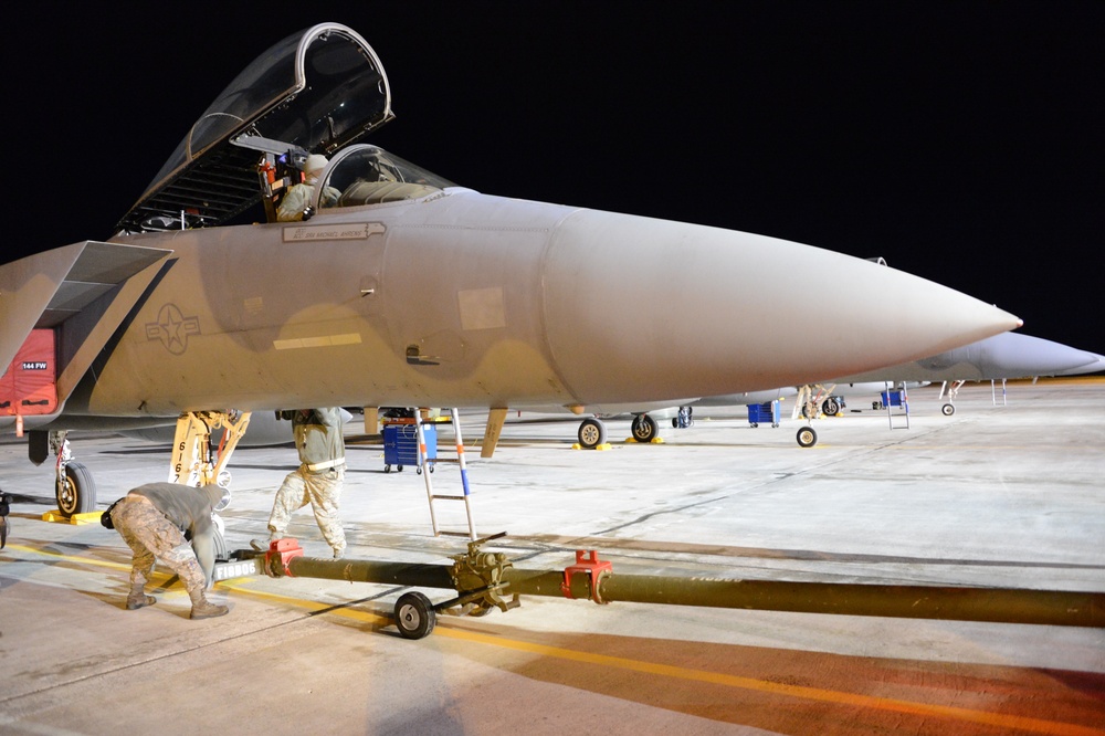 A night-time military flight line scene showing ground crew personnel performing maintenance or pre-flight operations on what appears to be an F-15 Eagle fighter jet. The aircraft's canopy is open and multiple technicians are working around the aircraft on a concrete tarmac. Additional aircraft and ground support equipment are visible in the background.