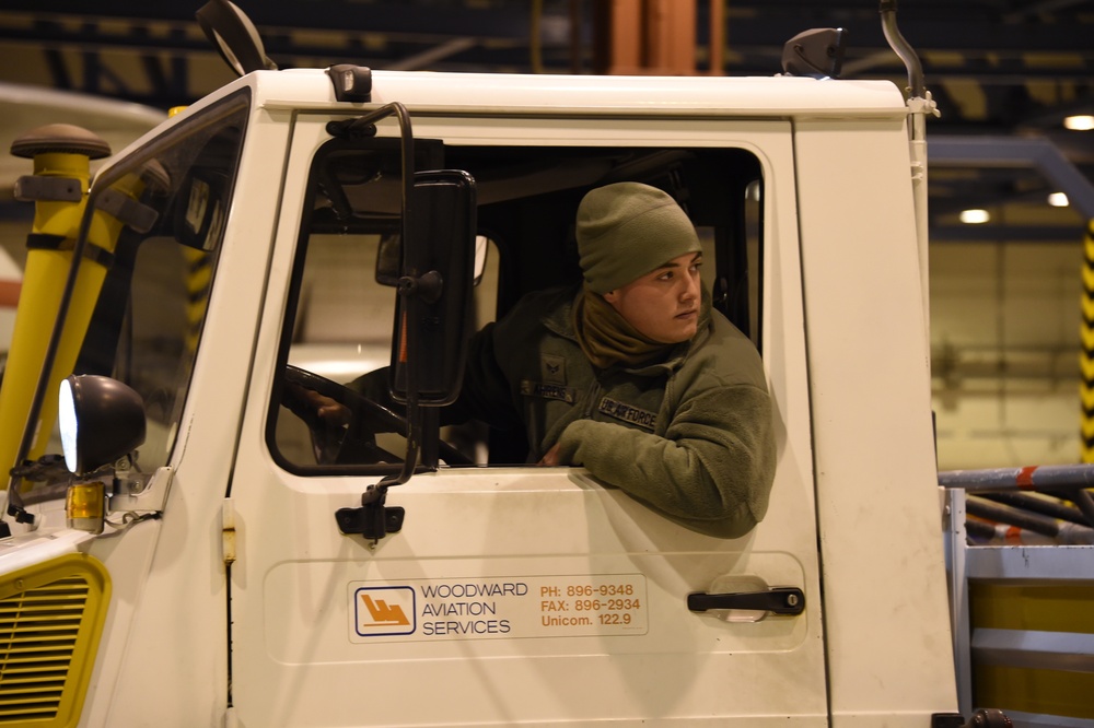 A person wearing U.S. Air Force fleece and an olive green beanie is seated in the cab of a Woodward Aviation Services ground support vehicle inside what appears to be an aircraft hangar. The individual is looking to the side, suggesting they are maneuvering or monitoring their surroundings while operating the vehicle. The vehicle's door panel displays Woodward Aviation Services contact information including phone, fax, and Unicom frequency.
